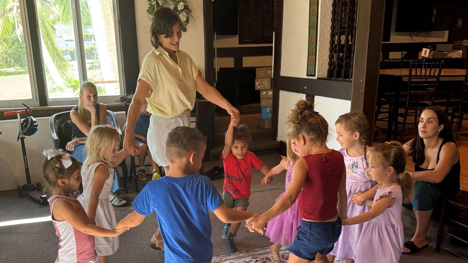 A teacher and a circle of young children holding hands during a Saturday class, with two parents watching from the side